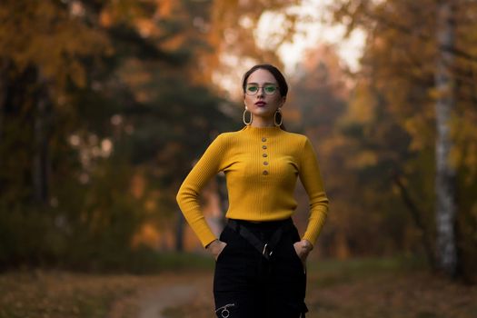 Young long haired brunette in yellow knitted jumper and eyeglasses looking at camera while standing against blurred foliage of autumn trees in park