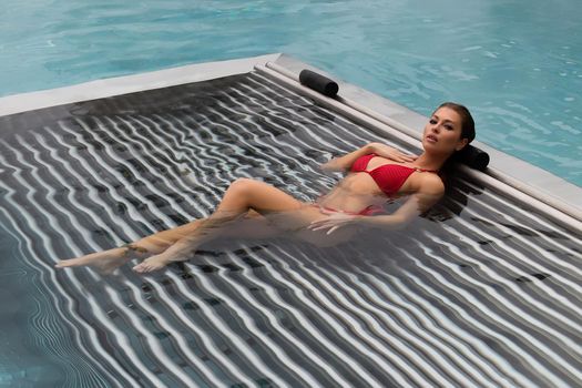 High angle of slim barefoot woman in red swimwear lying on grates in clean water of pool on resort