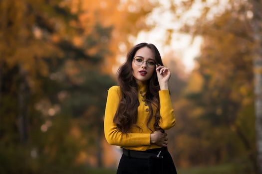 Young long haired brunette in yellow knitted jumper and eyeglasses looking at camera while standing against blurred foliage of autumn trees in park