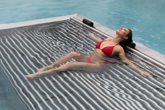 High angle of slim barefoot woman in red swimwear lying on grates in clean water of pool on resort