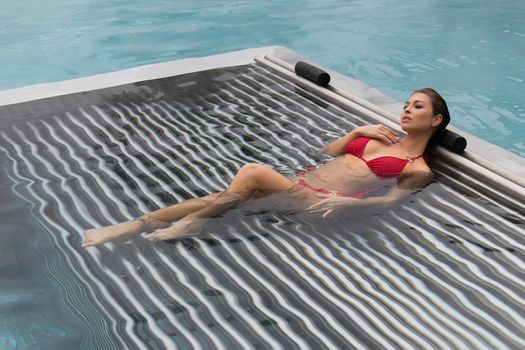 High angle of slim barefoot woman in red swimwear lying on grates in clean water of pool on resort