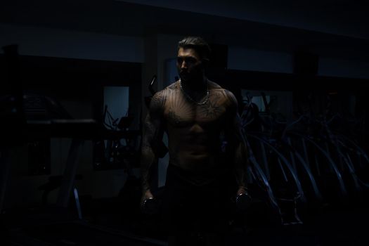 Muscular male athlete with tattooed naked torso standing in dark gym and doing dumbbell curls during workout