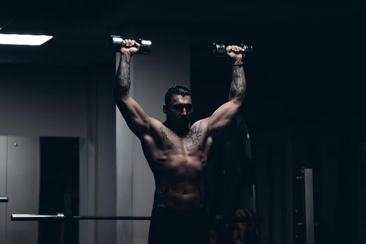 Muscular male athlete with tattooed naked torso standing in dark gym and doing dumbbell curls during workout