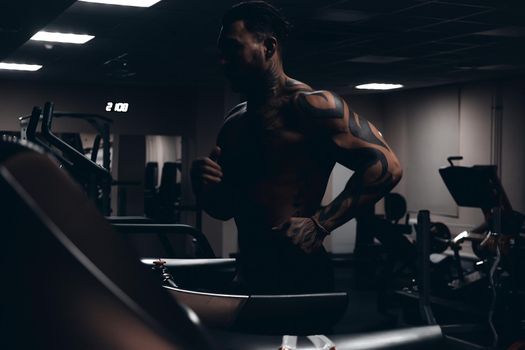 Muscular male athlete with tattooed naked torso standing in dark gym and doing dumbbell curls during workout