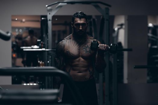 Muscular male athlete with tattooed naked torso standing in dark gym and doing dumbbell curls during workout