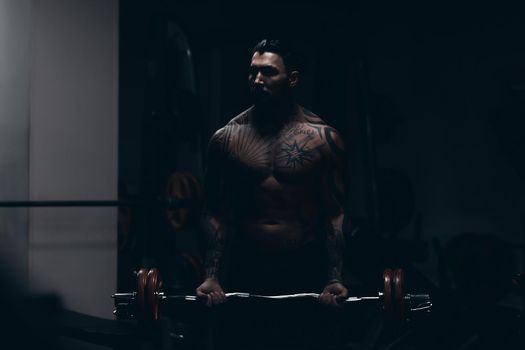 Muscular male athlete with tattooed naked torso standing in dark gym and doing dumbbell curls during workout