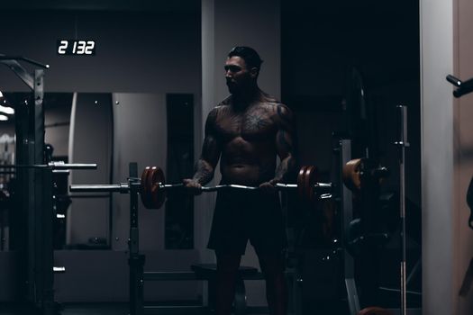 Muscular male athlete with tattooed naked torso standing in dark gym and doing dumbbell curls during workout