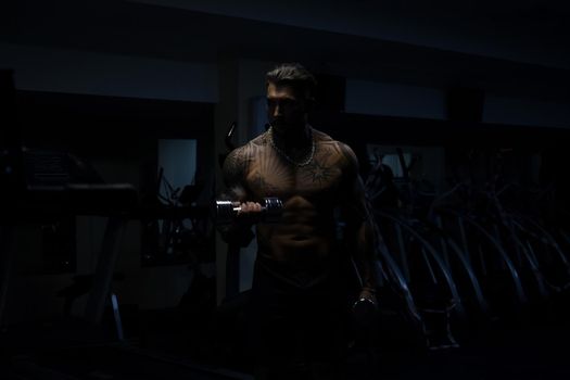 Muscular male athlete with tattooed naked torso standing in dark gym and doing dumbbell curls during workout