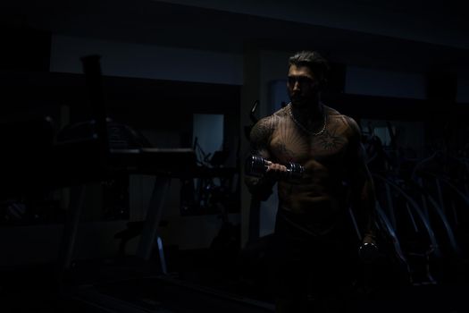 Muscular male athlete with tattooed naked torso standing in dark gym and doing dumbbell curls during workout