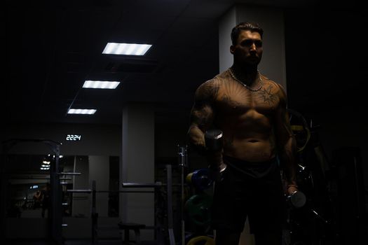 Muscular male athlete with tattooed naked torso standing in dark gym and doing dumbbell curls during workout