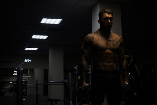 Muscular male athlete with tattooed naked torso standing in dark gym and doing dumbbell curls during workout