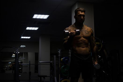 Muscular male athlete with tattooed naked torso standing in dark gym and doing dumbbell curls during workout