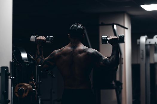 Muscular male athlete with tattooed naked torso standing in dark gym and doing dumbbell curls during workout