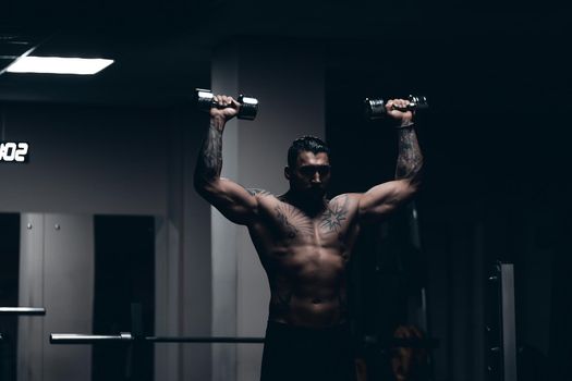 Muscular male athlete with tattooed naked torso standing in dark gym and doing dumbbell curls during workout