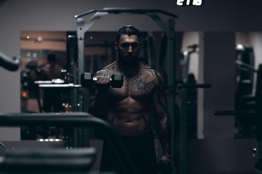 Muscular male athlete with tattooed naked torso standing in dark gym and doing dumbbell curls during workout