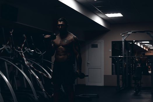 Muscular male athlete with tattooed naked torso standing in dark gym and doing dumbbell curls during workout