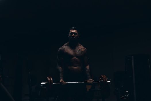 Muscular male athlete with tattooed naked torso standing in dark gym and doing dumbbell curls during workout