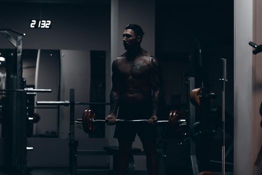 Muscular male athlete with tattooed naked torso standing in dark gym and doing dumbbell curls during workout