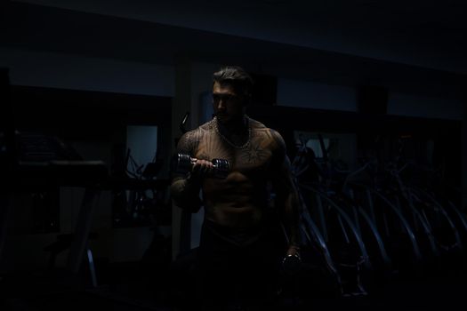 Muscular male athlete with tattooed naked torso standing in dark gym and doing dumbbell curls during workout