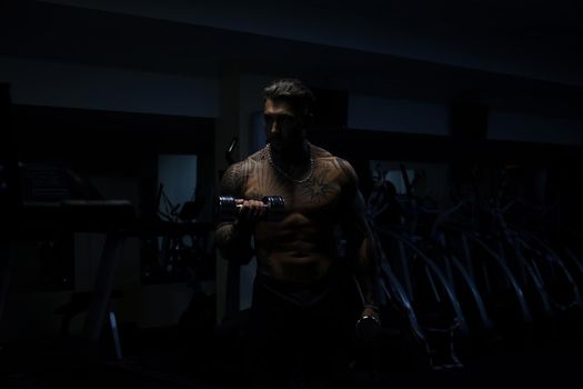 Muscular male athlete with tattooed naked torso standing in dark gym and doing dumbbell curls during workout