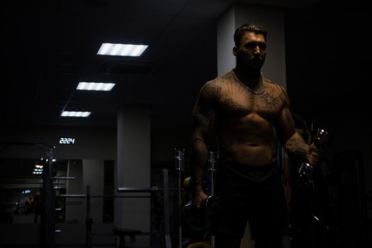 Muscular male athlete with tattooed naked torso standing in dark gym and doing dumbbell curls during workout