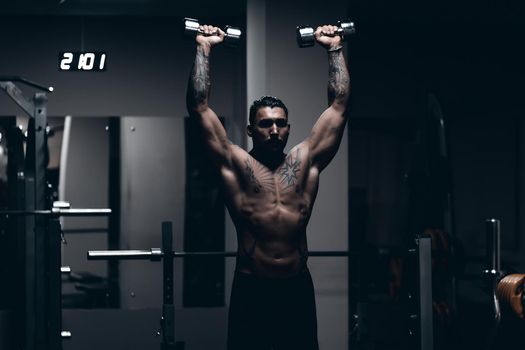 Muscular male athlete with tattooed naked torso standing in dark gym and doing dumbbell curls during workout