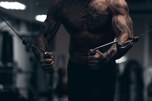 Muscular male athlete with tattooed naked torso standing in dark gym and doing dumbbell curls during workout