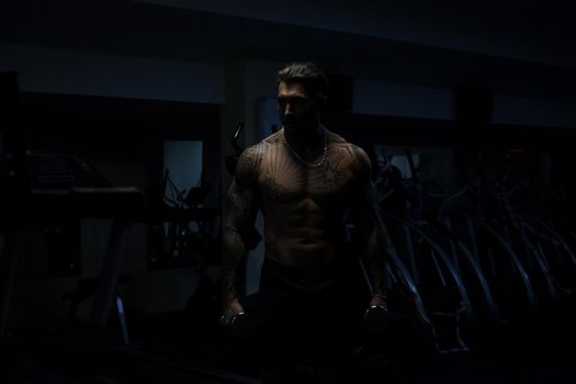 Muscular male athlete with tattooed naked torso standing in dark gym and doing dumbbell curls during workout