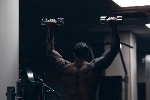 Muscular male athlete with tattooed naked torso standing in dark gym and doing dumbbell curls during workout