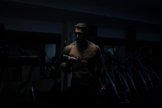 Muscular male athlete with tattooed naked torso standing in dark gym and doing dumbbell curls during workout