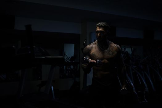 Muscular male athlete with tattooed naked torso standing in dark gym and doing dumbbell curls during workout