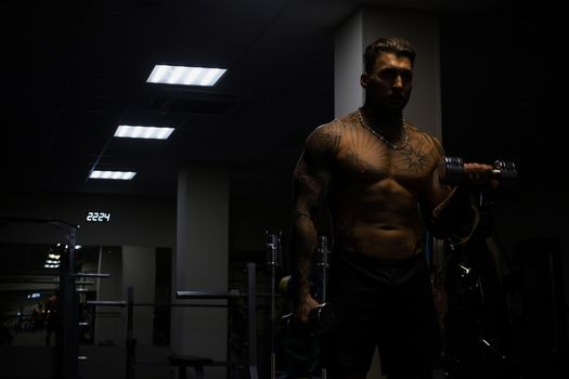 Muscular male athlete with tattooed naked torso standing in dark gym and doing dumbbell curls during workout