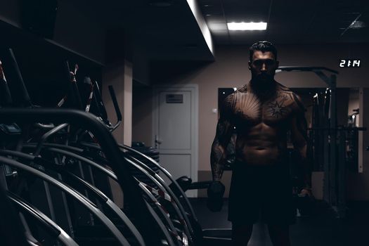 Muscular male athlete with tattooed naked torso standing in dark gym and doing dumbbell curls during workout