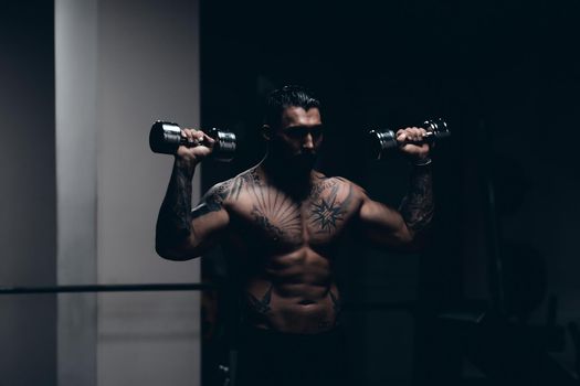 Muscular male athlete with tattooed naked torso standing in dark gym and doing dumbbell curls during workout