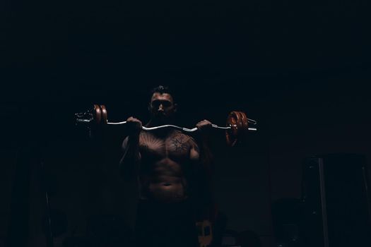 Muscular male athlete with tattooed naked torso standing in dark gym and doing dumbbell curls during workout