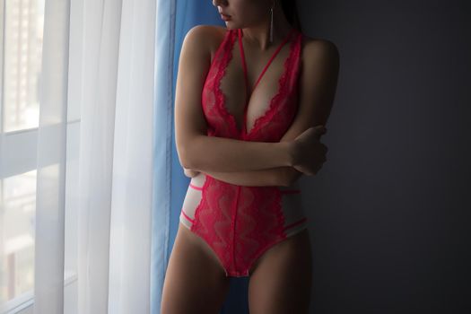 Unrecognizable female with perfect body in sexy red lace lingerie standing in dark bedroom and looking at window