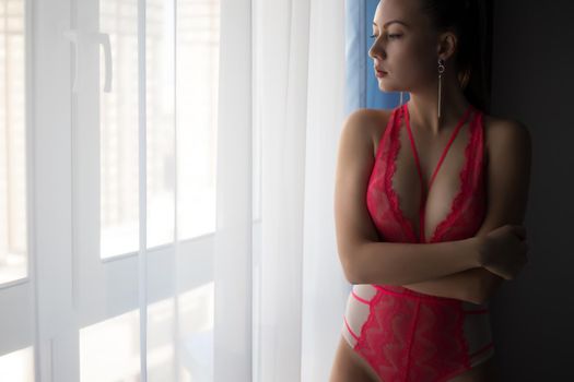 Unrecognizable female with perfect body in sexy red lace lingerie standing in dark bedroom and looking at window