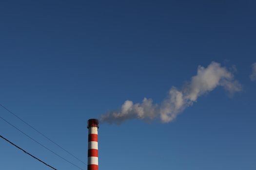 Red and white industrial chimney with smoke against blue sky background.