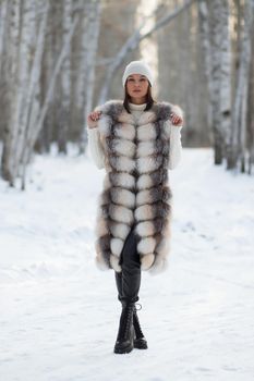 Gorgeous young brunette in fur vest and white knitwear looking confidently away against trees in winter woods