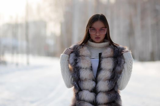 Gorgeous young brunette in fur vest and white knitwear looking confidently away against trees in winter woods