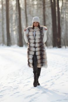 Gorgeous young brunette in fur vest and white knitwear looking confidently away against trees in winter woods