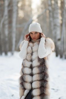 Gorgeous young brunette in fur vest and white knitwear looking confidently away against trees in winter woods