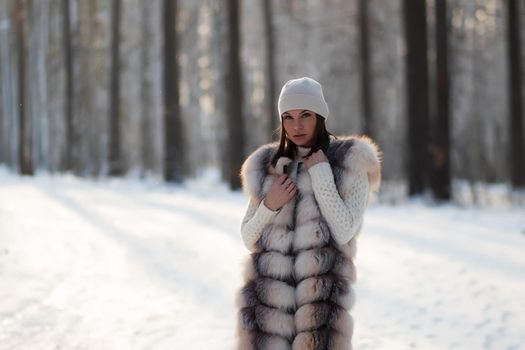 Gorgeous young brunette in fur vest and white knitwear looking confidently away against trees in winter woods