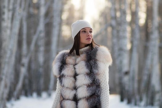 Gorgeous young brunette in fur vest and white knitwear looking confidently away against trees in winter woods