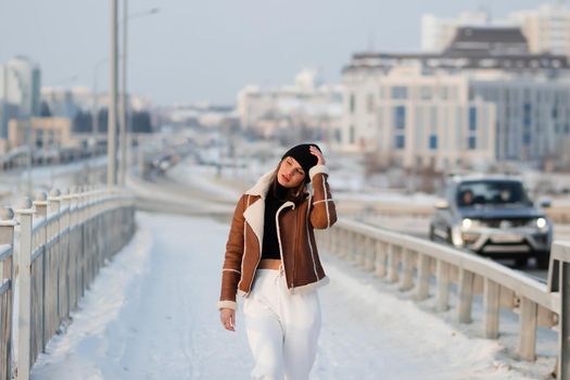 Alluring woman wearing brown sheepskin jacket with hat and looking at camera standing on snowy city bridge