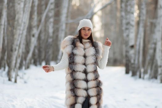 Gorgeous young brunette in fur vest and white knitwear looking confidently away against trees in winter woods