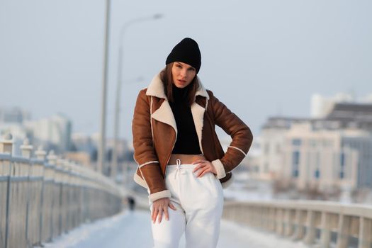 Alluring woman wearing brown sheepskin jacket with hat and looking at camera standing on snowy city bridge