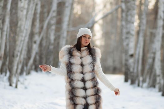 Gorgeous young brunette in fur vest and white knitwear looking confidently away against trees in winter woods