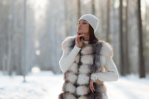 Gorgeous young brunette in fur vest and white knitwear looking confidently away against trees in winter woods