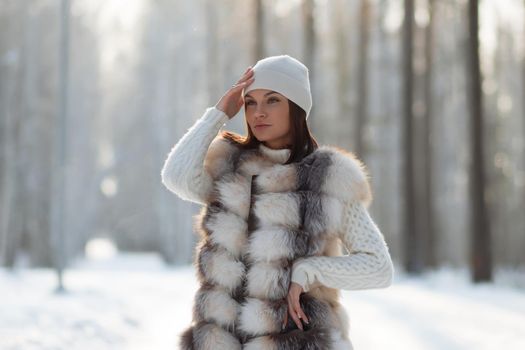 Gorgeous young brunette in fur vest and white knitwear looking confidently away against trees in winter woods